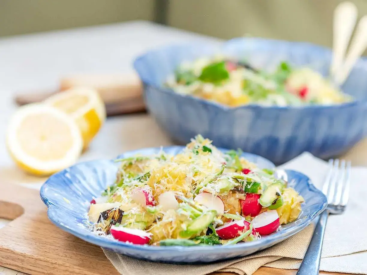 En härlig potatissallad med sparris, rädisor och hyvlad parmesanost. Serveras på en blå tallrik med citroner i bakgrunden.