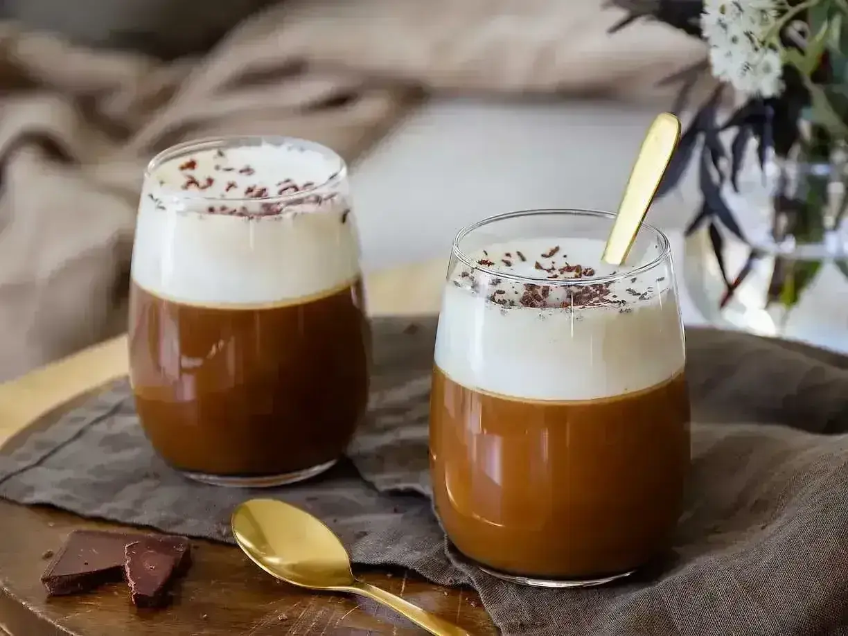 Två glas med krämig irish coffee, toppad med vispgrädde och chokladströssel. En gyllene sked vilar i det ena glaset.