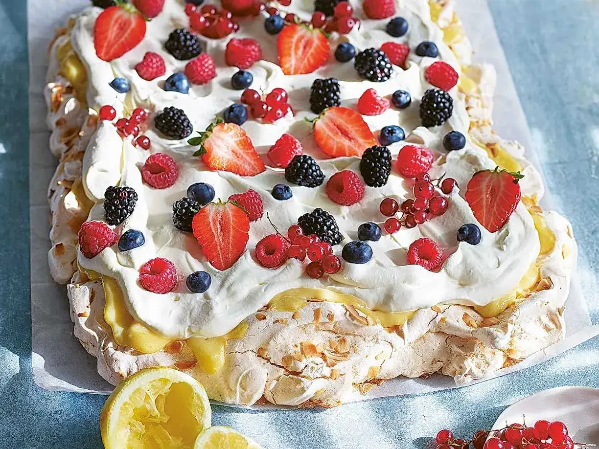 En stor pavlova långpanna täckt med vispgrädde, färska bär som jordgubbar, hallon, blåbär och vinbär, samt en gul lemon curd.