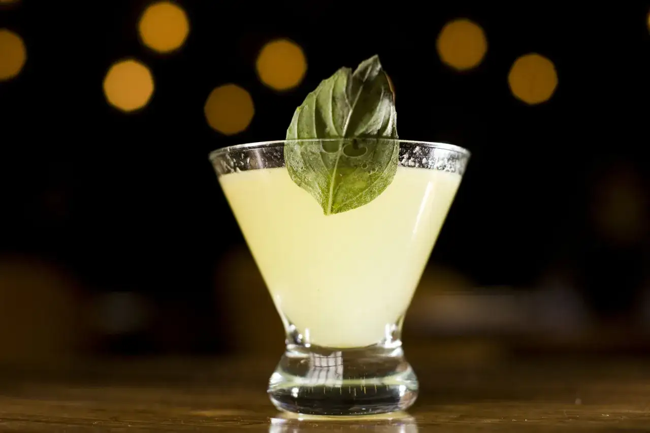 En ljusgul drink med en basilikakvist som garnityr, serverad i ett martiniglas mot en mörk bakgrund med bokeh-ljus.