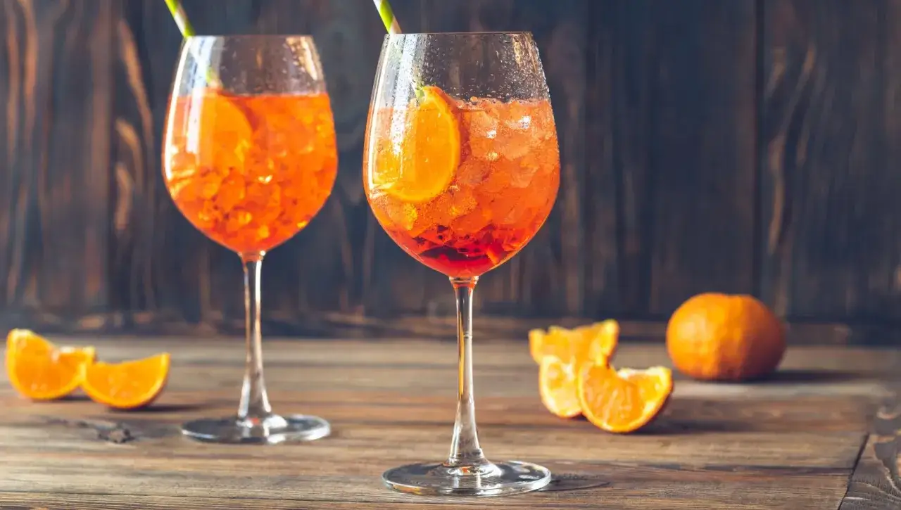 Två glas med Aperol Spritz, is och apelsinskivor, på ett träbord med apelsinklyftor och en hel apelsin.