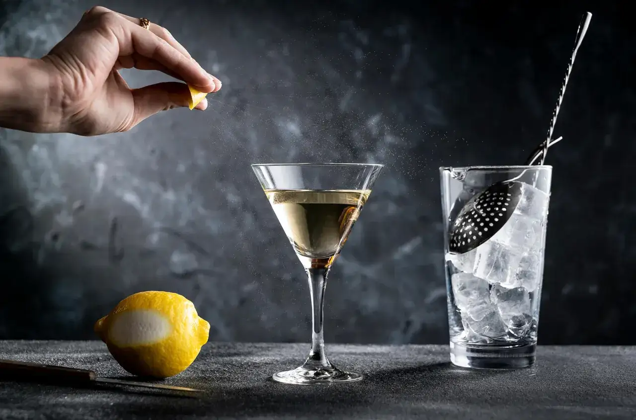 En hand pressar en citron över ett martiniglas, som om den förbereder en klassisk James Bond-drink. En citronskal och isbehållare syns bredvid.