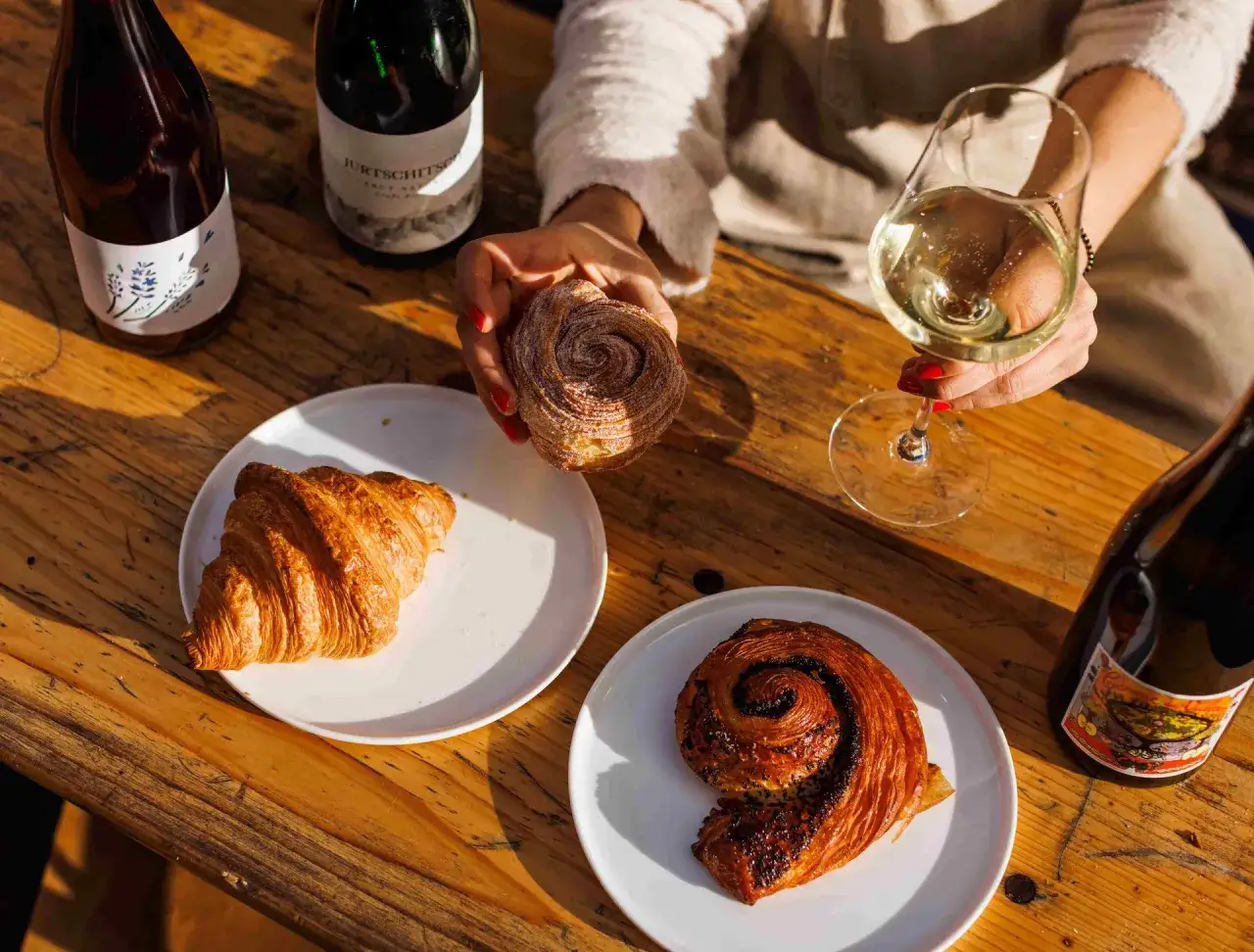 En hand håller en sockerdränkt bakelse bredvid en croissant och en chokladsnäcka. Flera flaskor vin, varav en märkt