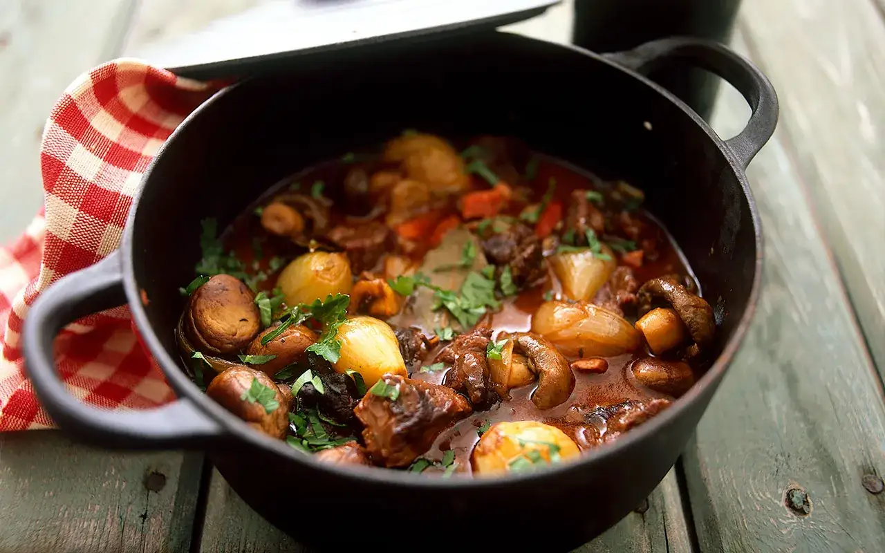 En mustig boeuf bourguignon i gjutjärnsgryta med champinjoner, lök och örter.