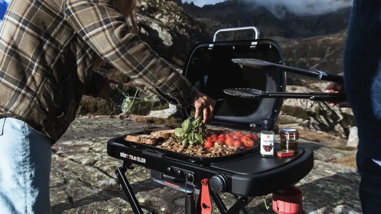 En person l&auml;gger f&auml;rska &ouml;rter p&aring; en weber liten grill med tomater och svamp i bergen.