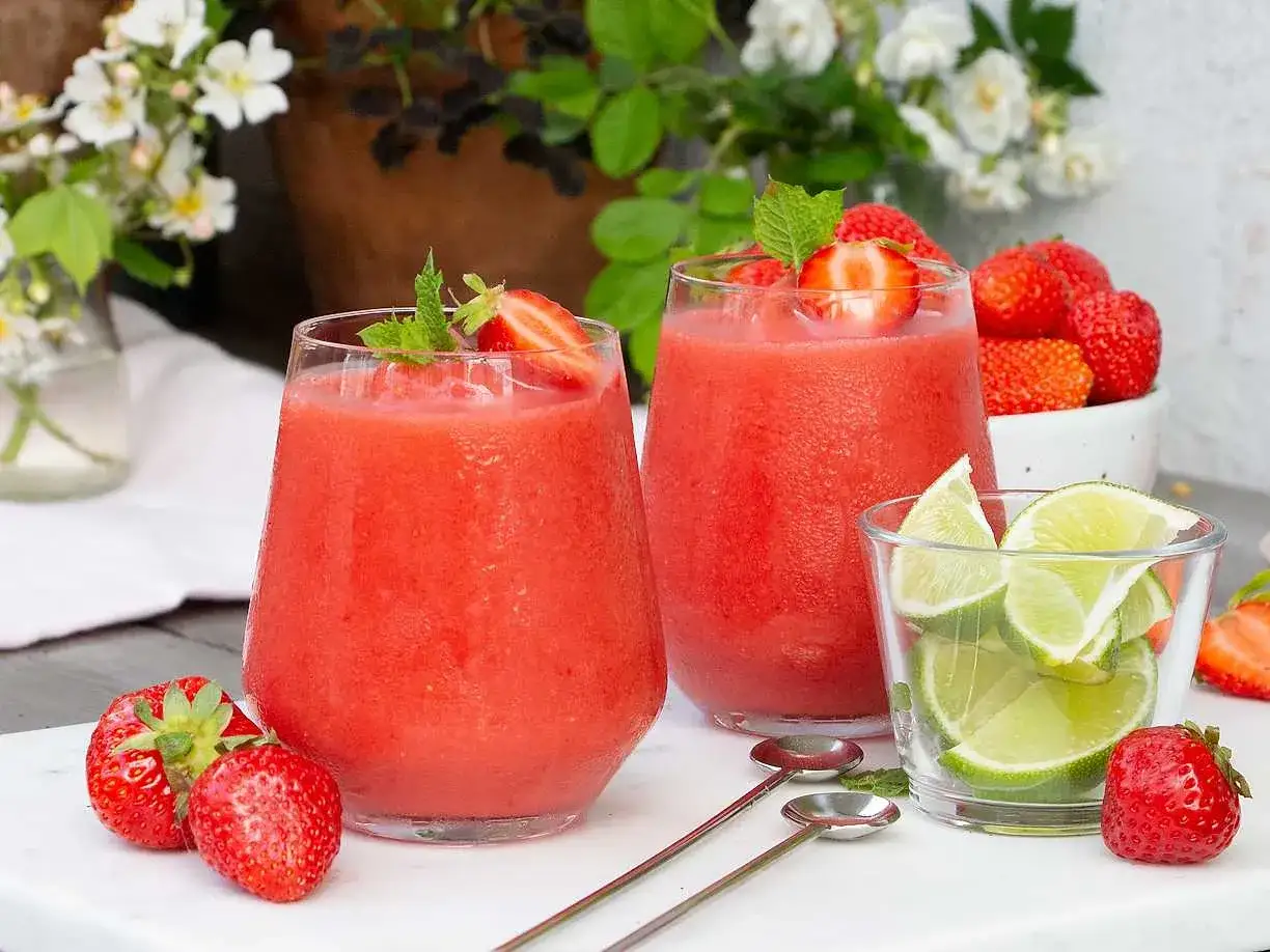 Två frostiga jordgubbsdaiquiri-drinkar med mynta och färska jordgubbar. En skål med limeklyftor och fler jordgubbar i bakgrunden.