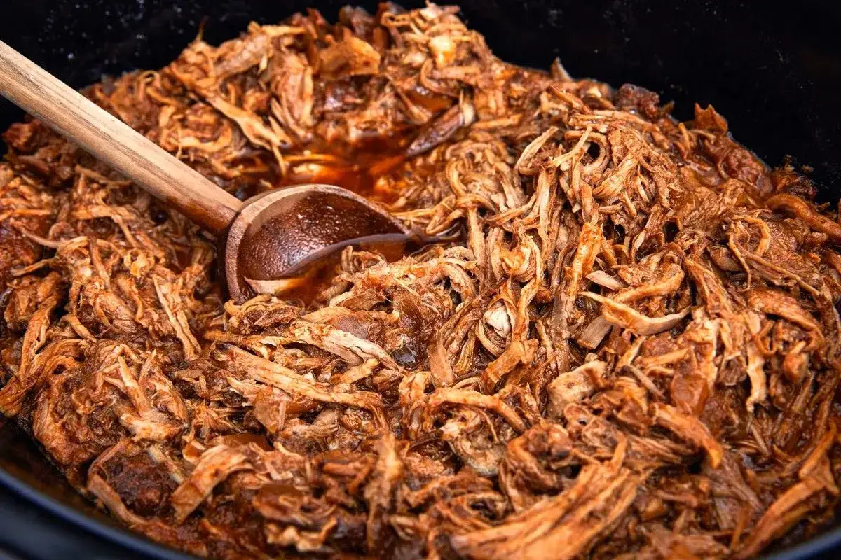 En stor gryta med saftig, l&aring;ngkokt pulled pork i en m&ouml;rk s&aring;s. En tr&auml;sked vilar i den l&auml;ckra pulled pork grytan.