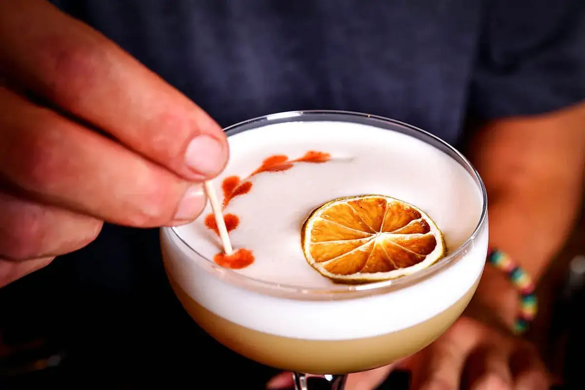 En hand dekorerar en perfekt whisky sour med en tunn pinne, en torkad lime och ett m&ouml;nster av bitters.