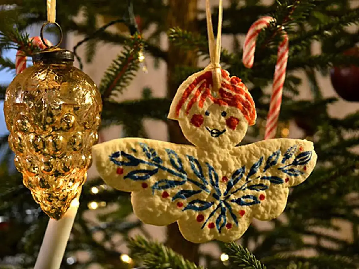 En ängel av vita pepparkakor hänger på julgranen, dekorerad med blåa blad och röda bär. Bredvid syns en guldfärgad julgranskula.
