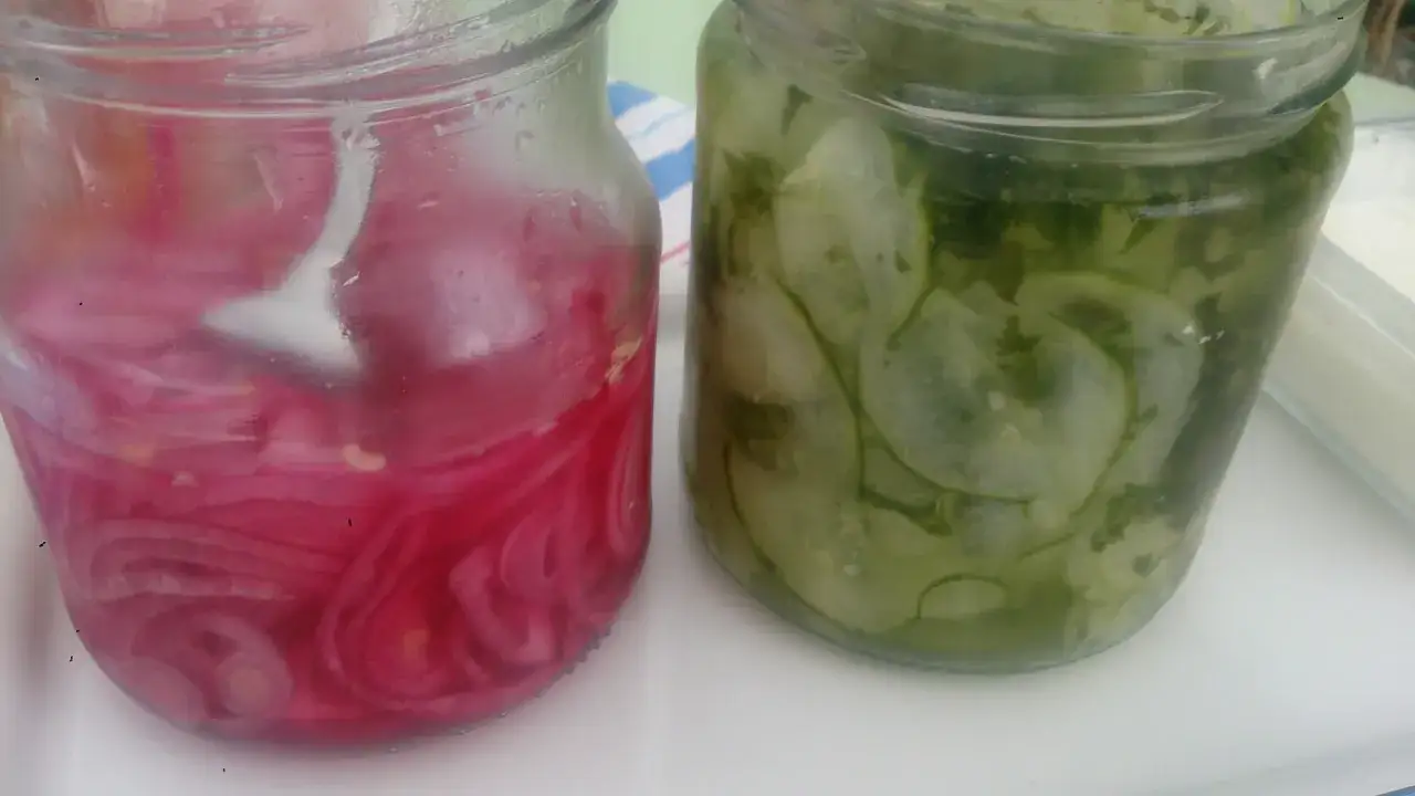 Två glasburkar med inlagd gurka och rödlök. Den ena burken innehåller skivad gurka med dill, den andra tunt skivad rödlök i en rosa lag.